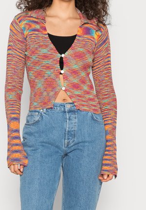 Femme porte un cardigan coloré, à manches longues, boutonné et court, par-dessus un haut noir et un jean taille haute bleu clair.
