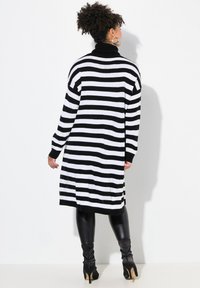 Cardigan lungo a righe bianche e nere con colletto alto, vestibilità rilassata, polsini a costine e spacco laterale, abbinato a leggings neri lucidi e stivale.