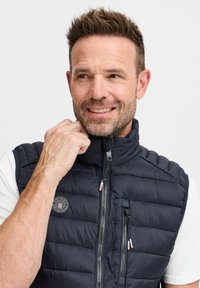 Navy padded vest with high collar, zippered front, and side pockets. Features a circular logo on the left chest. Lightweight fabric.