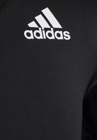 Nahaufnahme von schwarzem Stoff mit dem weißen Adidas-Logo, das drei diagonale Streifen und fetten Text auf einem Kleidungsstück zeigt.