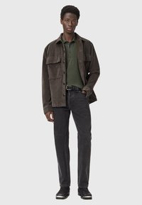 Mörkbrun kordflortjacka med två stora framfickor över en grön polo, tillsammans med svarta jeans och strukturerade svarta kängor.