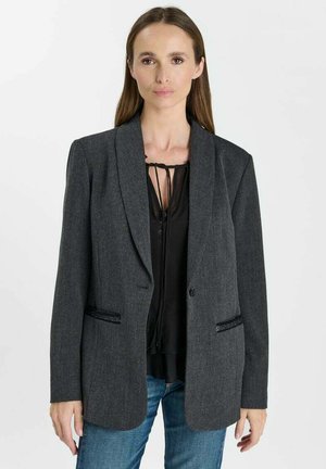 Frau, die einen dunkelgrauen Blazer über einer schwarzen Bluse mit Bindedetails und blauen Jeans trägt, steht vor einem schlichten hellen Hintergrund.