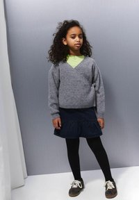 Siva, pletena ovita bluzica z dolgimi rokavi in stranskim vezanjem, v kombinaciji s temno denim midi krilom v plasteh in črnimi nogavicami, obuta v rjave superge.