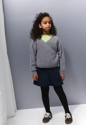 Szary, dziany sweter w kształcie narzuty z długimi rękawami i wiązaniem z boku, zestawiony z ciemnoniebieską, warstwową spódnicą i czarnymi rajstopami, noszony z brązowymi trampkami.