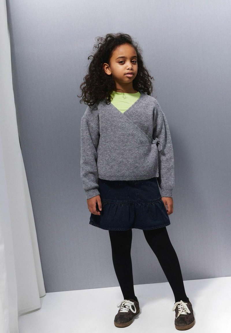 Siva, pletena ovita bluzica z dolgimi rokavi in stranskim vezanjem, v kombinaciji s temno denim midi krilom v plasteh in črnimi nogavicami, obuta v rjave superge.