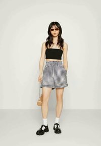 Top preto sem alças, calções xadrez de cintura alta, meias brancas, sapatos de plataforma pretos e uma pequena bolsa redonda. Modelo em pé contra um fundo liso.