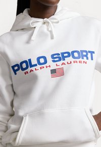 Personne portant un sweat à capuche blanc avec le texte « POLO SPORT RALPH LAUREN » et un petit drapeau américain imprimé sur le devant.