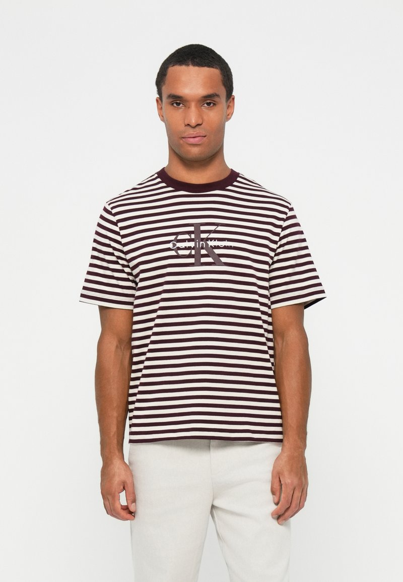 Dryžuota maroon ir kreminės spalvos marškinėliai su trumpomis rankovėmis ir apvaliu kaklu; centre yra Calvin Klein logotipas. Minkšta medvilnė.