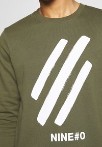 Olivgrön sweatshirt med en texturerad vit grafik av tre penseldrag och "NINE#0" tryckt nedan i ett djärvt, modernt teckensnitt.