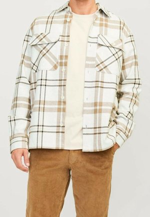 Mann trägt beige-weiß kariertes Hemd über einem cremefarbenen T-Shirt, kombiniert mit braunen Cordhosen, steht mit einer Hand in der Tasche.