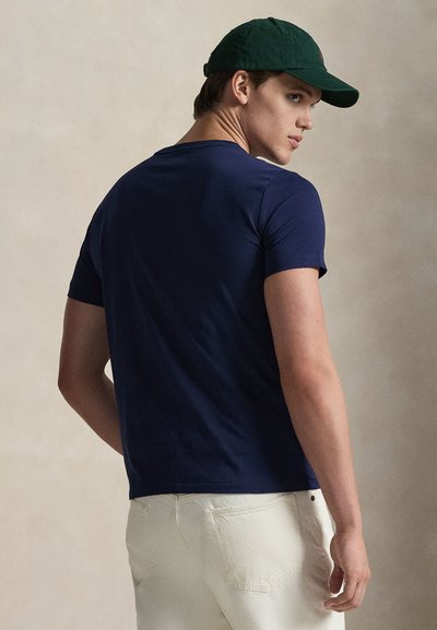 Μπλουζάκι από βαμβάκι σε σκούρο μπλε χρώμα με κοντά μανίκια, οπίσθια όψη. Συνδυασμένο με ανοιχτόχρωμο παντελόνι και πράσινο καπέλο. Ομαλή υφή, εφαρμοστή σχεδίαση.