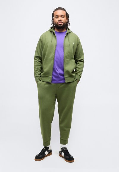 Άνδρας με dreadlocks, φοράει πράσινο φούτερ με φερμουάρ, παντελόνια φόρμας στο ίδιο χρώμα, μοβ μπλουζάκι, λευκές κάλτσες και μαύρα αθλητικά παπούτσια, στέκεται με τα χέρια στις τσέπες.