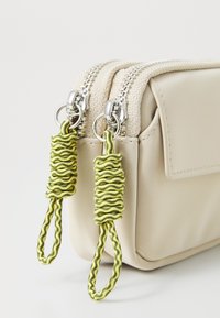 Sac zippé beige en matériau texturé, avec deux tirettes de fermeture éclair en métal argenté ornées de boucles en paracorde verte.