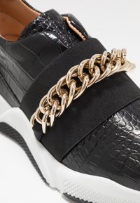 Baskets noires avec une tige à imprimé crocodile texturé, dotées d'une large sangle élastique noire et d'un accent en chaîne dorée. Semelle en caoutchouc blanc.