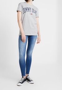 T-shirt in cotone grigio con stampa "TOMMY JEAN", abbinato a jeans in denim blu aderenti. Sneakers scure con lacci bianchi, stile casual.