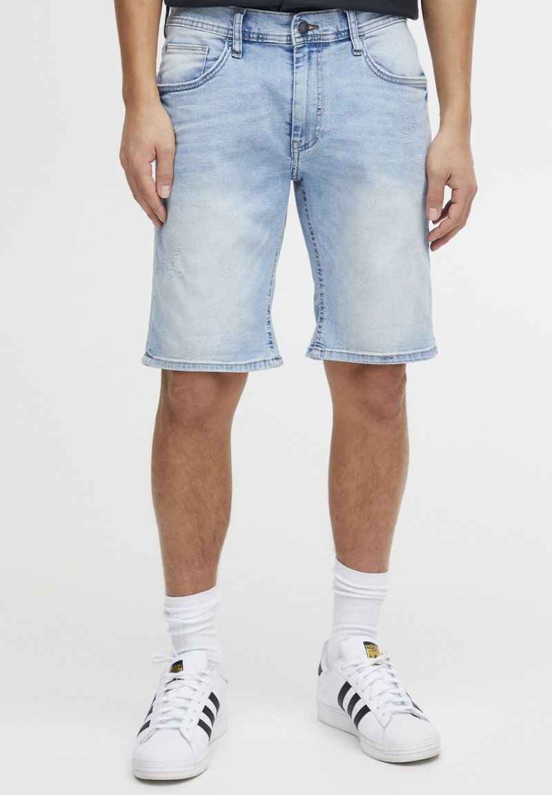 Shorts de mezclilla azul claro con un ajuste relajado, dobladillo deshilachado y un sutil detalle desgastado. Llevados con calcetines blancos y zapatillas negras a rayas.
