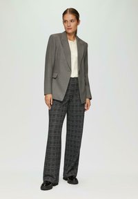 Grijze blazer met getailleerde schouders, gecombineerd met high-waisted geruite broek in zwart-wit. Glanzende zwarte schoenen maken de look compleet.