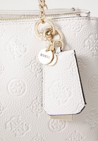 Witte handtas met textuur en een reliëflogo-patroon, gouden hardware en een afneembaar label met de merknaam "GUESS."