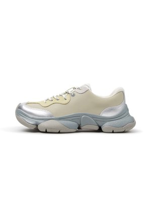 Deportiva gruesa en beige y plateado con suela gruesa y texturizada azul grisácea y cordones blancos, mostrada de lado sobre fondo blanco.
