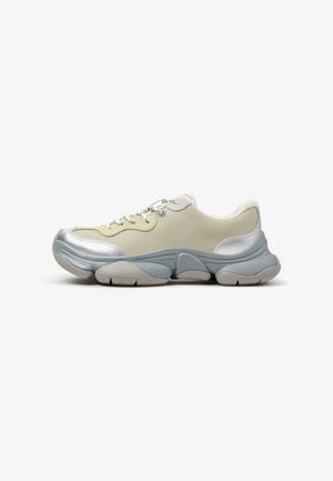 Deportiva gruesa en beige y plateado con suela gruesa y texturizada azul grisácea y cordones blancos, mostrada de lado sobre fondo blanco.