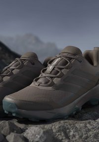Brązowe buty trekkingowe Adidas Terrex na skalistym terenie z teksturowaną podeszwą i siateczkową cholewką, na tle rozmytego górskiego krajobrazu.