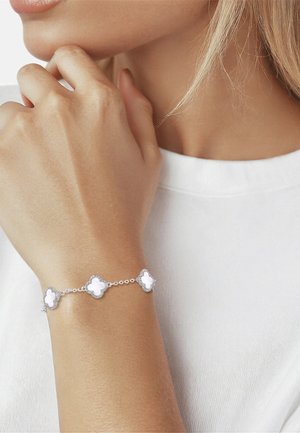 Pulsera de plata con charms blancos en forma de trébol de cuatro hojas vinculados, llevada en la muñeca de una persona con camisa blanca.