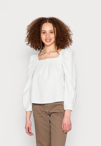 Blusa branca com riscas, de decote quadrado, mangas bufantes e um corte descontraído, combinada com calças castanhas. Textura suave, design casual.