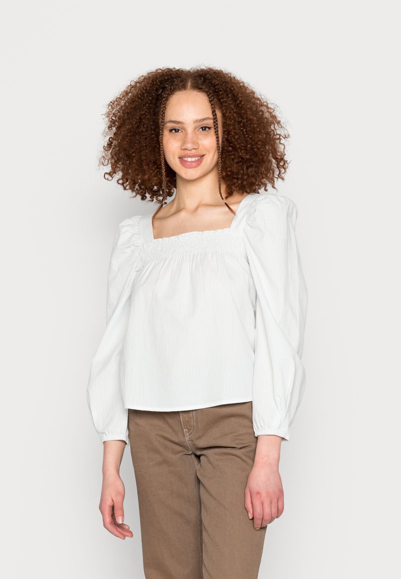 Blusa branca com riscas, de decote quadrado, mangas bufantes e um corte descontraído, combinada com calças castanhas. Textura suave, design casual.