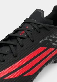 Μαύρο ποδοσφαιρικό παπούτσι με υφή στην επιφάνεια, μαύρα κορδόνια και ζωηρές κόκκινες διαγώνιες ρίγες στο πλάι.