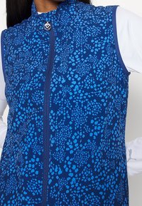 Blå mönstrad ärmlös zip-up topp som bärs över en vit långärmad tröja med en liten hjärtformad dragkedja.