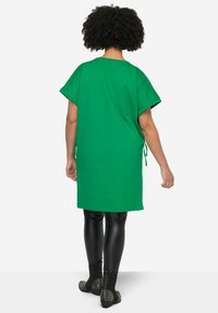 Túnica verde de gran tamaño con mangas cortas enrolladas, con lazos en los laterales. Combinada con leggings de imitación de cuero negros y botas de tobillo con tachuelas.