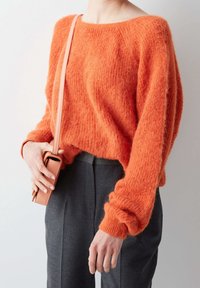 Suéter esponjoso de color naranja brillante con un escote amplio y mangas largas, combinado con pantalones gris con pliegues y sosteniendo una pequeña bolsa rosa.