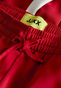 Raudoni sportiniai šortai su elastiniu juosmeniu, su balta virvele ir geltonu žymekliu "J.XX". Glotni, lengva medžiaga.