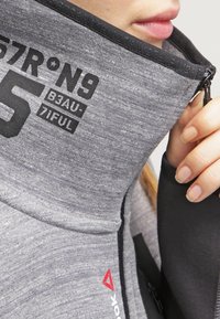 Giacca grigia con zip e colletto alto, caratterizzata da testo stampato in nero e dettagli con logo rosso. Il tessuto ha un aspetto morbido e testurizzato.