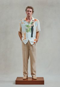 Korte mouwen overhemd met knoop in lichte kleuren met grafische ontwerpen, gecombineerd met beige broek en geweven instap schoenen, staande op een houten platform.