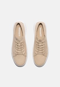 Beiga avslappnade sneakers gjorda av texturerat material, med vita snören, en gummisula och en liten logodetalj på sidan.