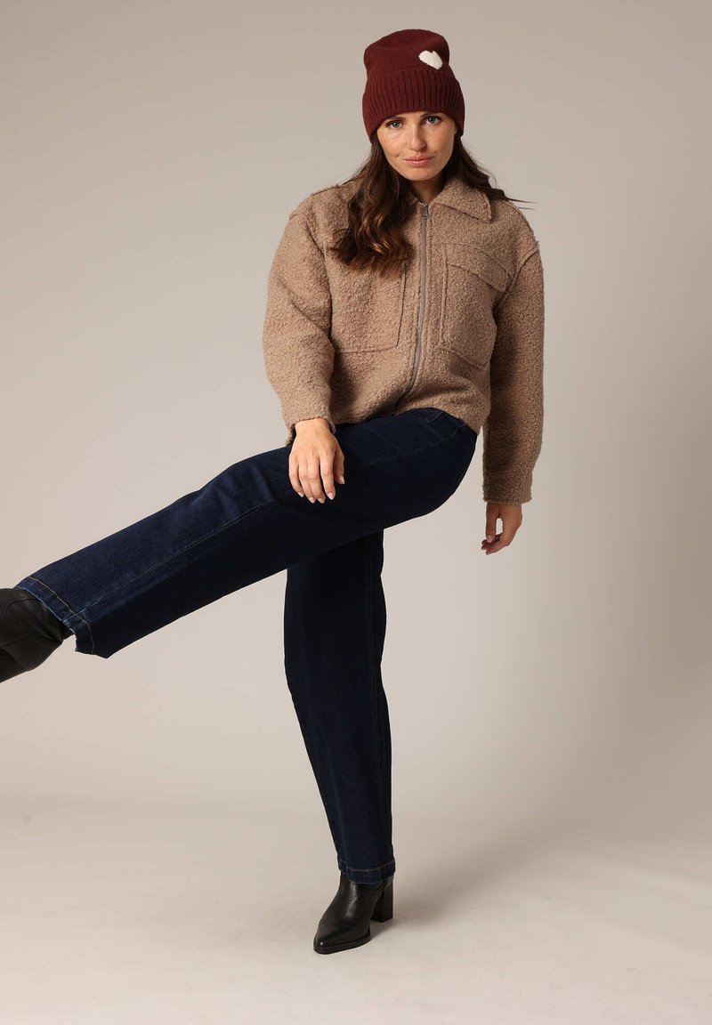 Veste zippée en fausse fourrure beige avec deux poches poitrine, associée à un jean droit bleu marine et des bottines noires. Bonnet en tricot bordeaux avec un cœur.