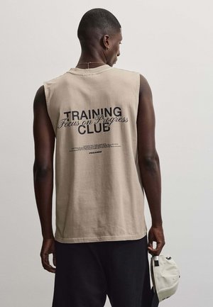Homme de dos, portant un débardeur beige avec "TRAINING CLUB Focus on Progress" imprimé dans le dos, tenant une casquette claire à la main.