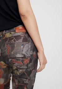 Pantaloni in denim a fantasia floreale in tonalità scure, dotati di tasche posteriori e etichetta in pelle del marchio. Tessuto testurizzato con accenti vivaci.