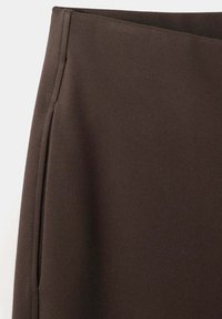 Gros plan d’un tissu brun foncé avec des coutures visibles le long d’une couture sur le côté gauche sur un fond clair.