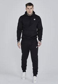 Black hoodie and sweatpants set, cotton blend, featuring a front pocket, ribbed cuffs, and a logo accent on the chest and thigh. White sneakers.