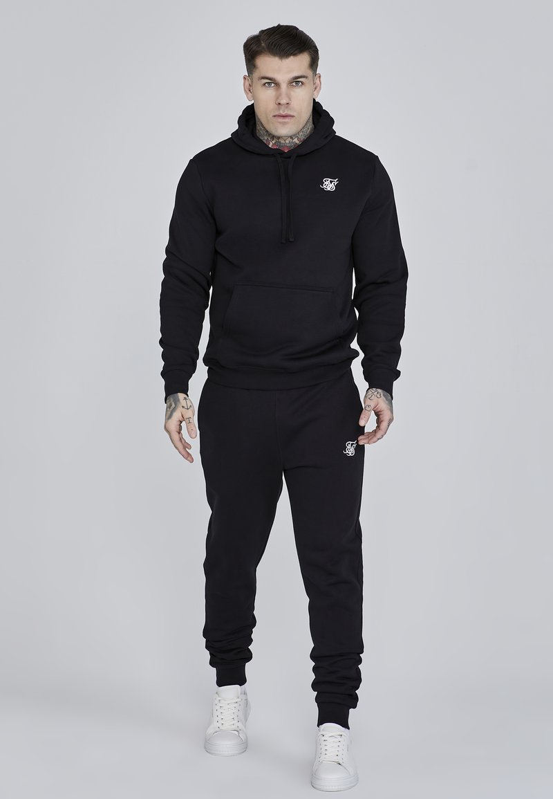 Black hoodie and sweatpants set, cotton blend, featuring a front pocket, ribbed cuffs, and a logo accent on the chest and thigh. White sneakers.