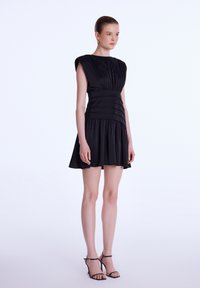 Robe noire avec un corsage ajusté, des plis, des manches courtes et une jupe évasée. Accompagnée de talons noirs à lanières.