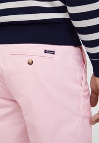 Lichtroze katoenen broek met een achterzak met een knoop en een marineblauw label. Gerealiseerd met een marineblauw en wit gestreept truitje.