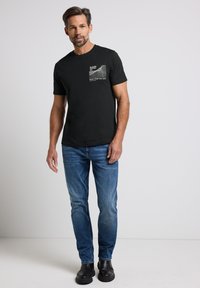 Camiseta de algodón negro con mangas cortas, que presenta una impresión gráfica y texto en el lado izquierdo del pecho; combinada con jeans de mezclilla azules y calzado negro.
