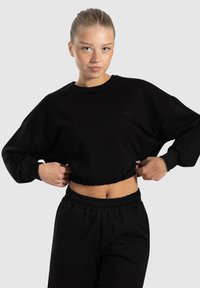 Schwarzer, cropped Sweatshirt mit langen Ärmeln, gerippten Bündchen und gesticktem Logo. Kombiniert mit schwarzen, elastischen Hosen mit Bund. Glatte Baumwolltextur.