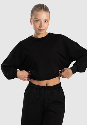 Sort cropped sweatshirt med lange ærmer, ribbede manchetter og broderet logo. Parret med sorte bukser med elastisk talje. Glat bomuldsstruktur.