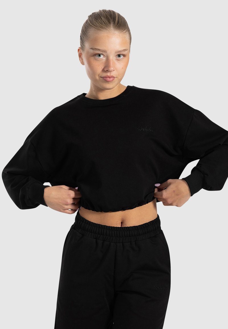 Schwarzer, cropped Sweatshirt mit langen Ärmeln, gerippten Bündchen und gesticktem Logo. Kombiniert mit schwarzen, elastischen Hosen mit Bund. Glatte Baumwolltextur.