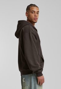 Sudadera marrón oscuro con un bolsillo canguro, corte oversize y puños de canalé. Mostrada en un modelo de lado contra un fondo liso.