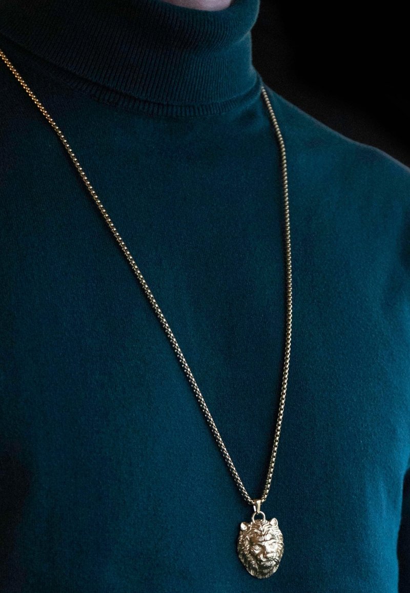 Pendentif en tête de lion doré sur une chaîne argentée, porté sur un col roulé vert foncé. La chaîne est texturée et le pendentif présente des détails fins.